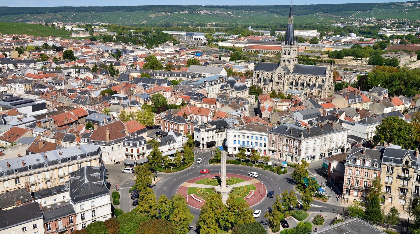 Place centrale animée d'une ville moyenne française avec commerces de proximité, terrasses et passants, baignée de lumière naturelle.